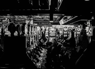 A dimly lit casino filled with rows of slot machines, some occupied by people. The machines' lights contrast with the dark surroundings, creating a vibrant yet subdued atmosphere.