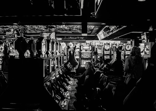 A dimly lit casino filled with rows of slot machines, some occupied by people. The machines' lights contrast with the dark surroundings, creating a vibrant yet subdued atmosphere.