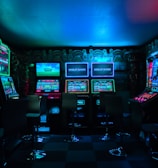 Dimly lit gaming arcade featuring a variety of brightly illuminated slot machines and video gaming screens. Neon signs and colorful displays create a vibrant atmosphere. The arrangement of gaming machines suggests an inviting space for entertainment, with chairs positioned for players.