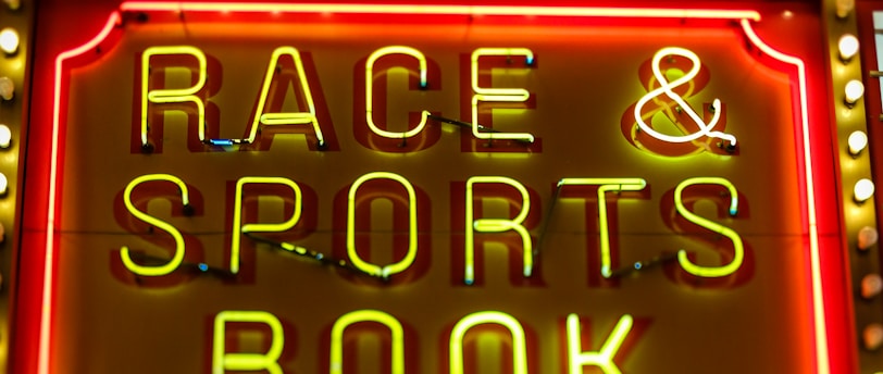Bright neon lights on a marquee sign display the words 'RACE & SPORTS BOOK'. The sign is surrounded by rows of yellow and white lights against a red background, evoking a vibrant, energetic atmosphere typically associated with casinos or entertainment venues.