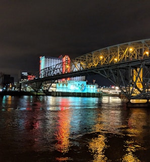 A brightly lit casino with neon signs and red lights is reflected in a river. A bridge stretches across the water, with an urban skyline visible in the background against an overcast night sky.