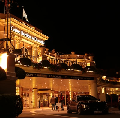 A luxurious casino at night, illuminated with warm, golden lights. The building features elegant architecture with columns and decorative elements. A few people are walking towards the entrance, and there are a couple of parked cars nearby.