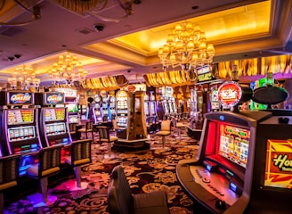 A vibrant casino interior filled with rows of colorful slot machines, each illuminated with bright lights and displays. The ceiling is adorned with elegant chandeliers and golden drapes, creating a luxurious atmosphere. The patterned carpet adds to the elaborate design of the space.