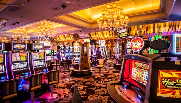 A vibrant casino interior filled with rows of colorful slot machines, each illuminated with bright lights and displays. The ceiling is adorned with elegant chandeliers and golden drapes, creating a luxurious atmosphere. The patterned carpet adds to the elaborate design of the space.