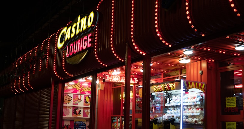 A brightly lit casino lounge entrance adorned with neon lights. The exterior features colorful signage including the words 'Casino' and 'Lounge' in vivid colors, with additional lights framing the building. Inside, a glimpse of a claw machine filled with plush toys and arcade games is visible. The area exudes an inviting yet bustling atmosphere typical of entertainment venues.
