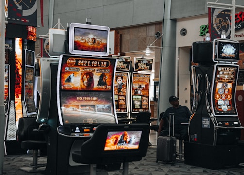 A collection of slot machines is situated inside a building with bright, colorful screens displaying various games and gaming themes. A person is seated nearby, seemingly relaxed with luggage beside them, and the atmosphere is lively with electronic displays.