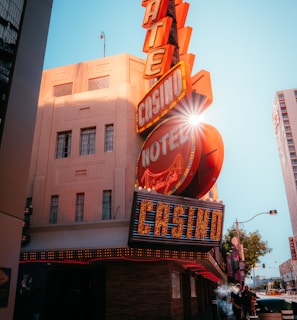 A vibrant city scene featuring a tall, illuminated casino sign with retro-style neon lights. The sign dominates the building facade with the words 'CASINO' and 'HOTEL'. Sunlight peeks through, creating a dramatic lens flare effect. The street is lively, with pedestrians casually walking and a hint of urban hustle.