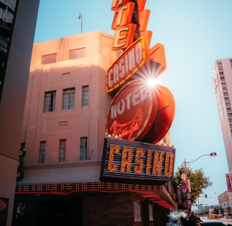 A vibrant city scene featuring a tall, illuminated casino sign with retro-style neon lights. The sign dominates the building facade with the words 'CASINO' and 'HOTEL'. Sunlight peeks through, creating a dramatic lens flare effect. The street is lively, with pedestrians casually walking and a hint of urban hustle.
